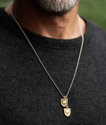 Man wearing the Courageous Love Tags necklace with two 14k gold vermeil shield-and-heart pendants on a silver chain, shown against a dark knit sweater.
